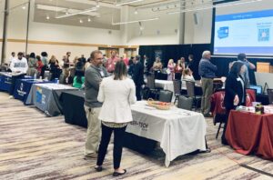 Groups of people in a large room visiting tables where information is being distributed.