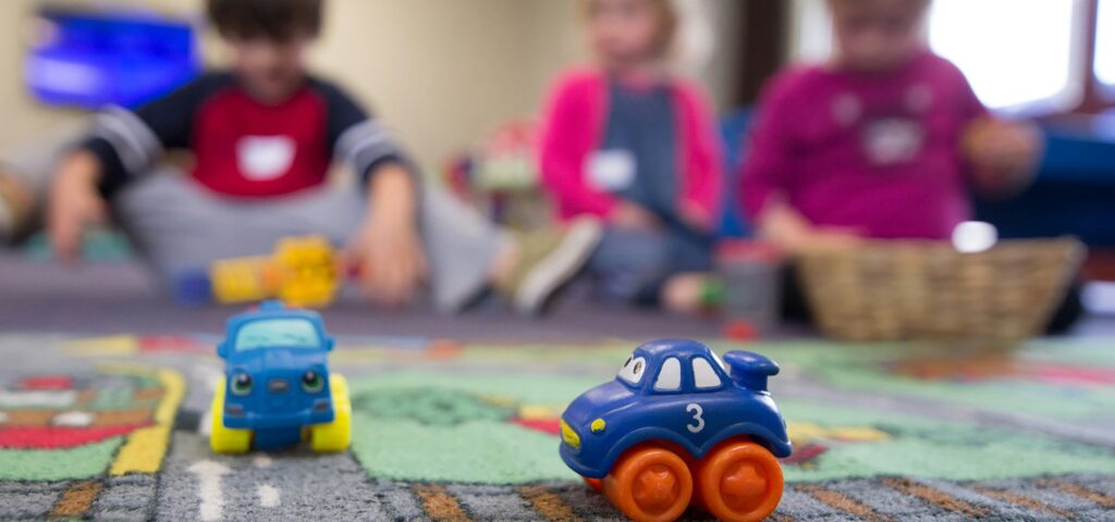 Toy cars in foreground of children playing on floor of classroom.