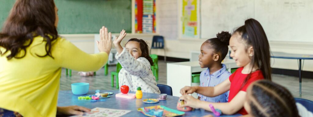 A teacher high-fives one of several children sitting at a classroom table.