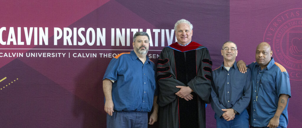 Four men stand in front of a banner for the Calvin Prison Initiative. Two on the left and one on the right are wearing blue uniforms. The man second from right is wearing academic robes.