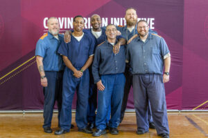 Six men stand in front of a banner for the Calvin Prison Initiative. They are wearing blue uniforms. 