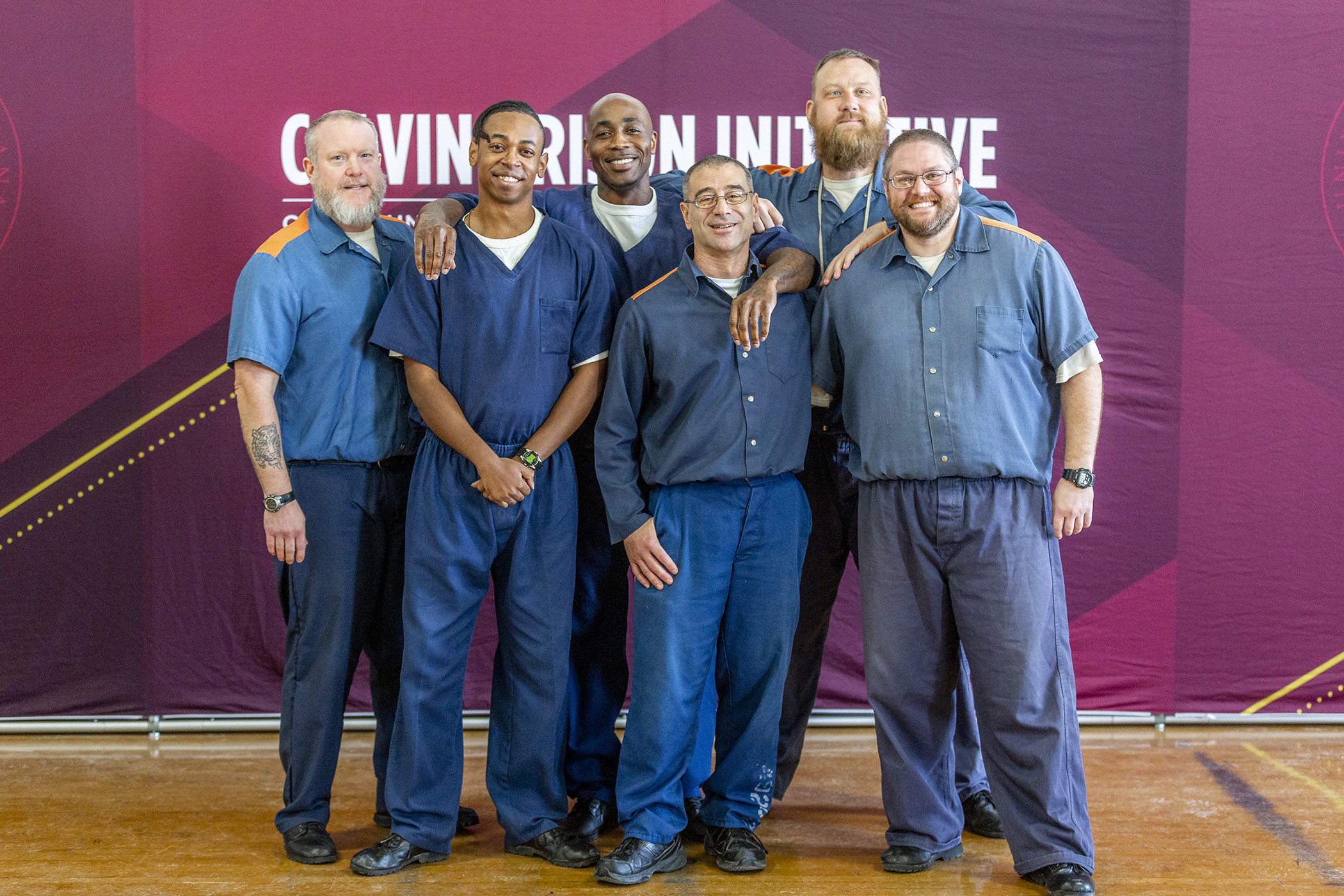 Six men stand in front of a banner for the Calvin Prison Initiative. They are wearing blue uniforms.