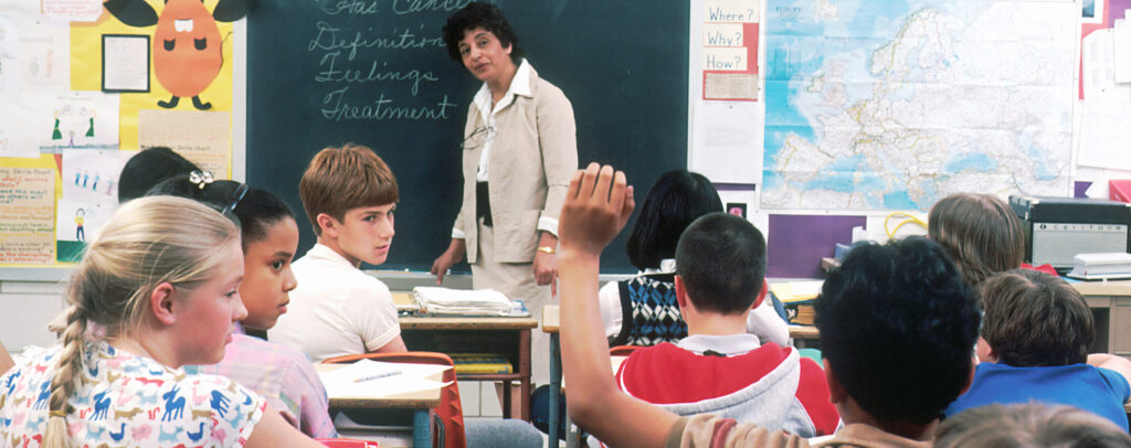 A teacher stands at a chalkboard at the front of classroom with several students. One of them is raising a hand.