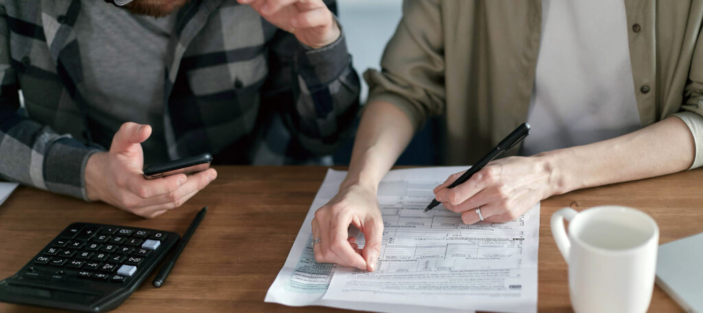 A man, left, and woman, faces not visible, work with a calculator and paper on a table.