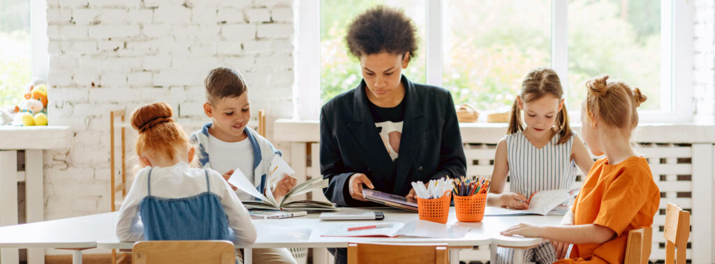 A teacher sits at a table with four children reading books.