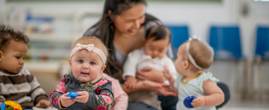 An infant in the foreground, with a caregiver and other toddler-aged children.