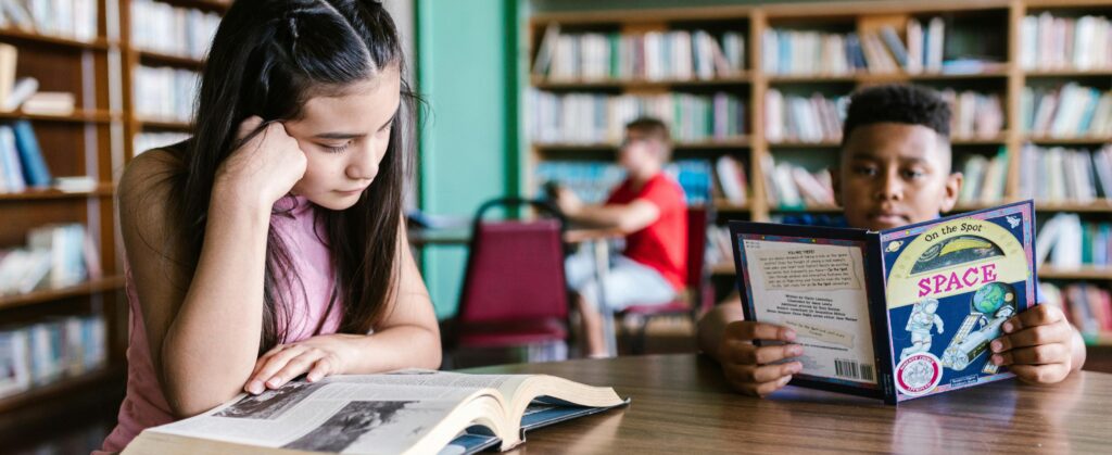 Two young students in foreground are reading books at a table in a library. Another student is in the background.