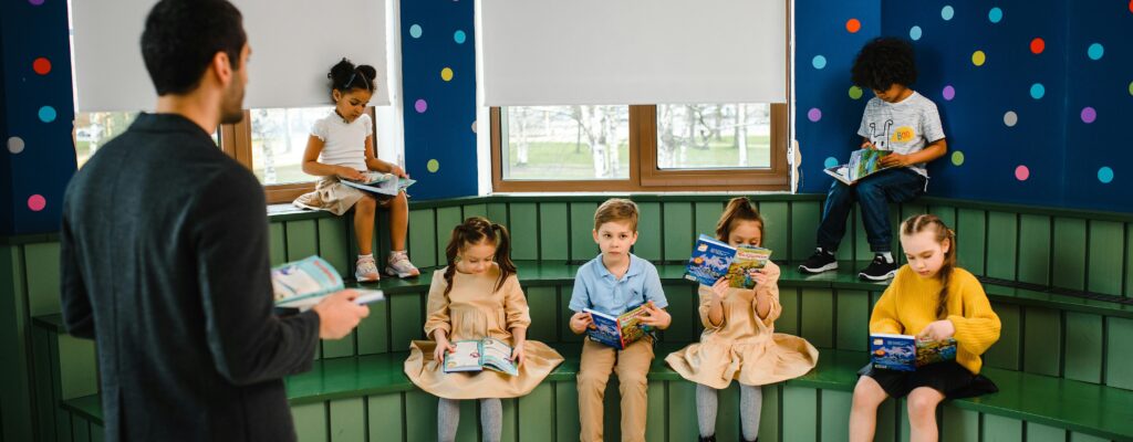 Children with books seated on risers, teacher at front.